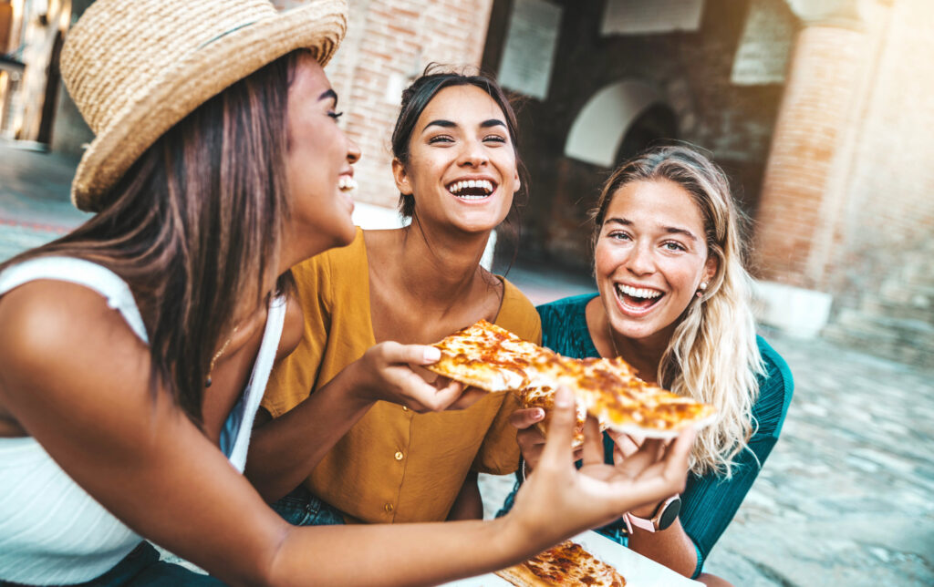 Three young female friends eating pizza sitting outside - Multiracial women enjoying street food in the city - Italian food culture and summer vacations concept - Christian Counselors of Mooresville Eating with friends makes campus life a lot more joyful.